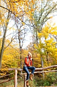 Blonde woman wearing gray puffer jacket sitting on wooden fence in forest