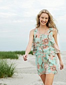Portrait of cheerful blonde woman in floral pattern summer dress walking on beach, smiling