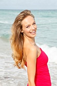 Portrait of beautiful blonde woman wearing red dress standing on beach, smiling