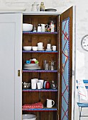 Open, wooden kitchen cupboard with crockery on shelves decorated with pretty trim