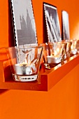 Lit tea light candles in glasses on orange shelf