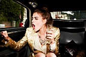 Excited woman wearing shiny golden dress holding glass of champagne while sitting in car