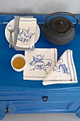 Teekanne, Teeschälchen & mit verschiedenen Motiven bestickte Leinenservietten auf blauer Kommode