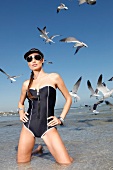 Sexy woman in black swimsuit, kneeling in water, seagulls in background