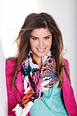 Pretty dark haired woman in pink jacket and colourful scarf leaning on wall, smiling