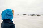 Contemplative blonde woman wearing coat and blue cap on beach, smiling
