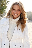 Pretty blonde woman wearing white jacket over white sweater standing on beach, smiling