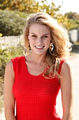 Blonde Frau in rotem Kleid am Strand, blickt lächelnd in die Kamera