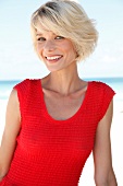 Portrait of pretty blonde woman with short hair wearing red top sitting on beach, smiling