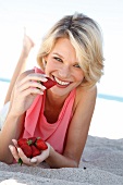 Blonde woman wearing pink top lying on sand while having strawberry