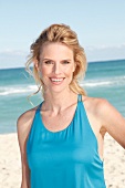 Portrait of pretty blonde woman wearing turquoise top standing on beach, smiling