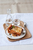 Lasagne al forno on plate with flask and glass of water on wooden serving tray