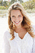 Portrait of beautiful blonde woman wearing white tunic blouse standing on beach, smiling