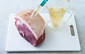 Pork with syringe and glass of drink on chopping board