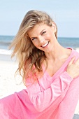 Portrait of pretty blonde woman wearing pink sweater sitting on beach, smiling