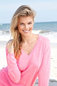 Portrait of pretty blonde woman wearing pink sweater sitting on beach, smiling