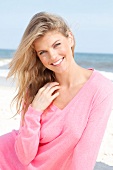 Portrait of pretty blonde woman wearing pink sweater sitting on beach, smiling