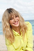 Portrait of cheerful blonde woman wearing yellow shirt sitting on beach, smiling