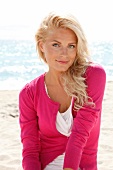 blonde Frau im Pulli in pink, lächelt in Kamera, am Strand