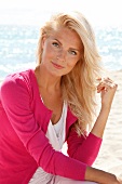 blonde Frau im Pulli in pink, lächelt in Kamera, am Strand