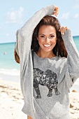 Pretty dark haired woman in gray sweatshirt with animal motif standing on beach, smiling