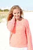 Blonde woman in an orange sweater on the beach, smiling at the camera