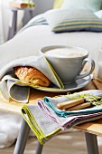 Pain au chocolat covered with a fabric napkin next to a cappuccino with a bundle of napkins on a wooden chair in front of a bed