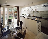 Modern, open kitchen with white countert and shell chairs made of dark wood around a glass top dining table and a wall of patio doors