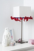A white table lamp decorated with red feather ribbon and vases decorated with flowers