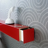 A white telephone on red shelf with an open drawer mounted on a wall hung with a geometric pattern