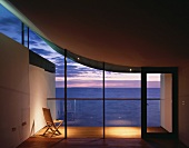 Stuhl auf beleuchteter Terrasse vor leerem Wohnraum mit Blick auf Abendhimmel unter gebogenem, einseitig schwebendem Betondach