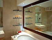Glass shelves and ornaments reflected in room-width mirror with wooden frame in stone-tiled bathroom