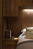 Bett vor holzverkleideter Wand mit indirekter Beleuchtung und Hängeschränkchen aus Holz