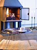 Gemütliche Winterstimmung mit brennendem Feuer im Kaminofen; davor ein runder Tabletttisch mit einer Tasse Kaffee und einem Buch