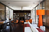 Offener Ess- und Wohnraum mit orangefarbener Stehlampe vor raumhohen Fenstern mit geschlossener Jalousie