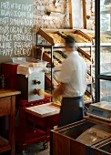 Verkaufsgestell mit selbst produzierten Nudeln und Mann in Aktion an Pasta-Maschine unter Kreidetafel mit handgeschriebenem Text