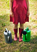 Frau mit roten Gummistiefeln steht zwischen zwei Giesskannen