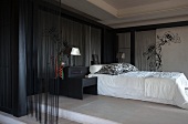 Elegant bedroom with black bamboo canes and thread curtains against walls around bed with white covers