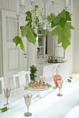 Silver wine goblets on white table cloth and candle chandelier decorated with plane tree leaves in white, country-house-style room