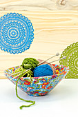 A wooden bowl covered with colorful paper with a knitted pattern; inside balls of wool and knitting needles