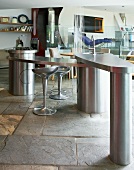 Stainless steel, freeform, designer kitchen with stone floor and silver retro bar stools at kitchen counter