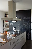 Küchenblock mit Dunstabzug in moderner Küche; dahinter eine wandhohe schwarze Schiefertafel