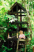 Doll on wooden ladder of a tree house surrounded by green leaves