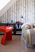 Dog lying on bed with striped blanket, red plastic chair and birch forest wallpaper in bedroom