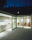 Innenhof mit weissen Bodenfliesen und Blick durch offene Terrassenschiebetüren in beleuchtete Küche und Chillecke
