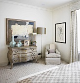 Light bedroom with Rococo chest of drawers next to light grey upholstered armchair
