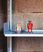 A Playmobil figure next to a retro day calendar on an aluminium shelf against a brick wall