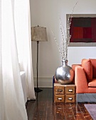 Silberfarbene Vase auf niedrigem Kästchen aus Holz und Stehlampe in Wohnzimmerecke