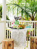 Filigraner Tisch und Hocker aus Baumstamm auf Veranda mit weiss lackiertem Geländer und Blick in tropischen Garten