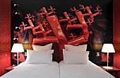 Knallrotes Hotelzimmer mit skurrilem Wandmotiv hinter blütenweissen Kopfkissen und brennenden Nachttischlampen